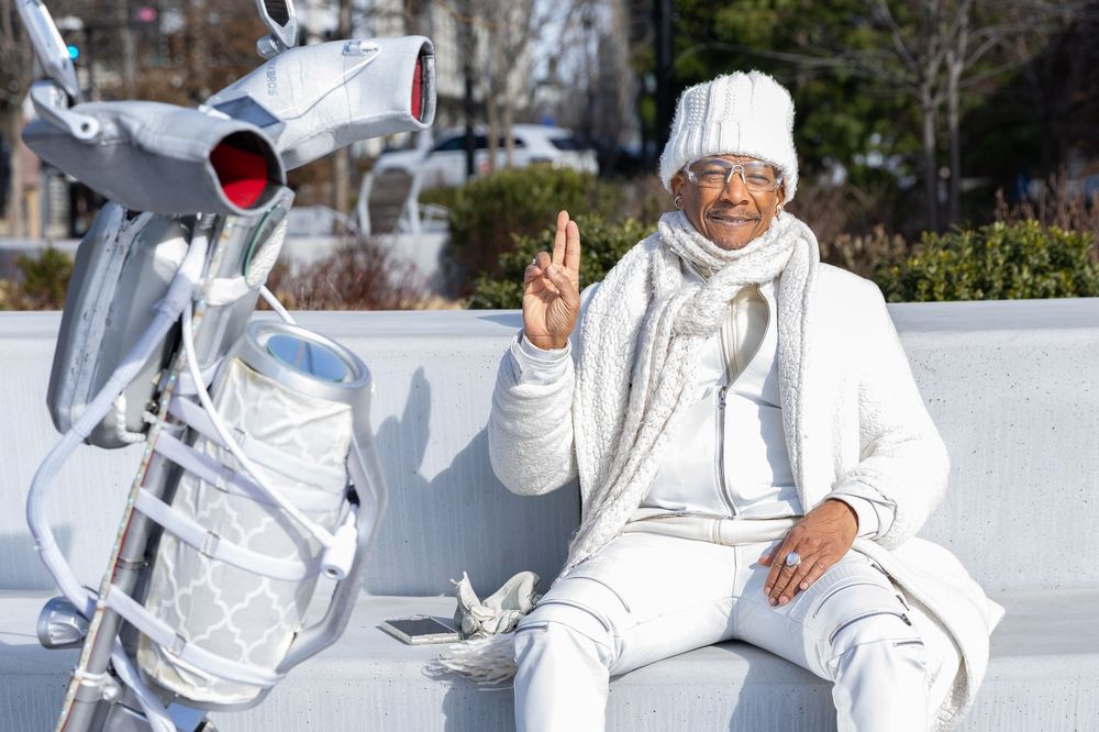 A person dressed in white winter clothing sits on a wall next to a silver scooter making a peace sign.