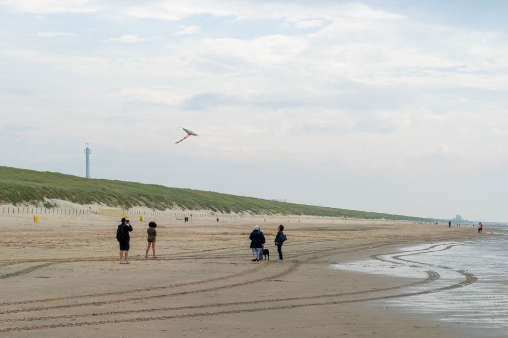 People walk along a peaceful beach shoreline with sand dunes and distant cliffs on a cloudy day.