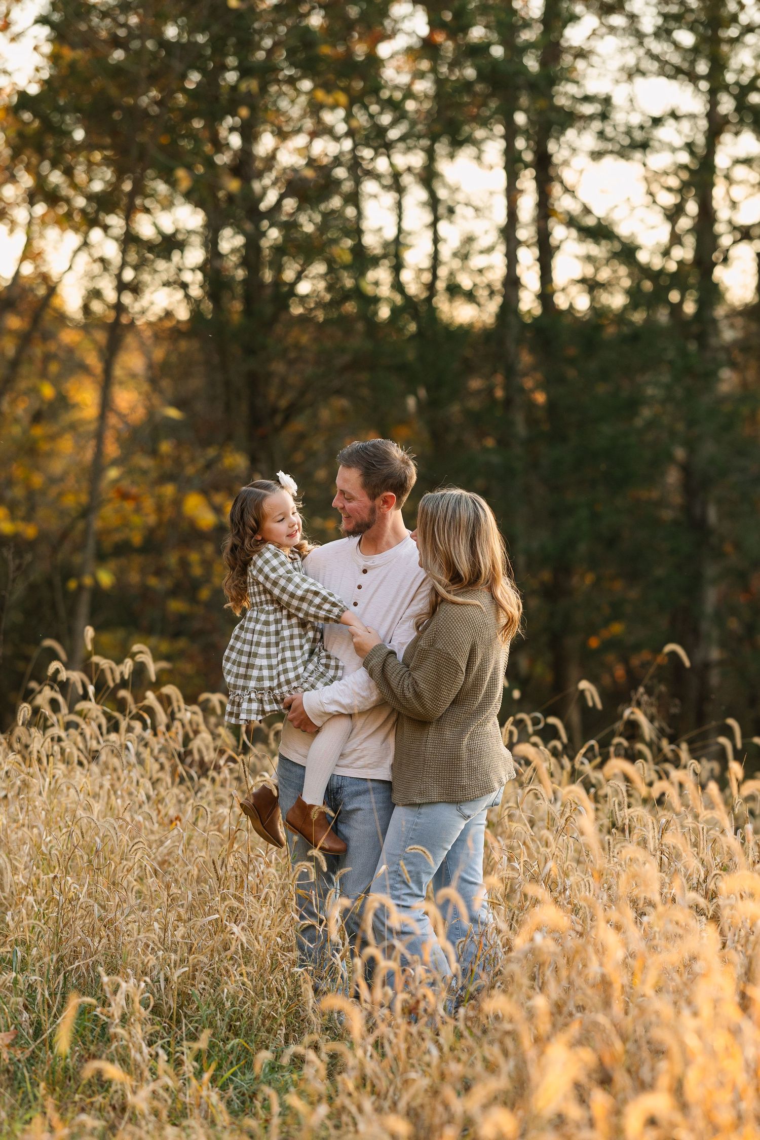 A family shares a tender moment together in a golden wheat field at sunset with autumn trees in the background.