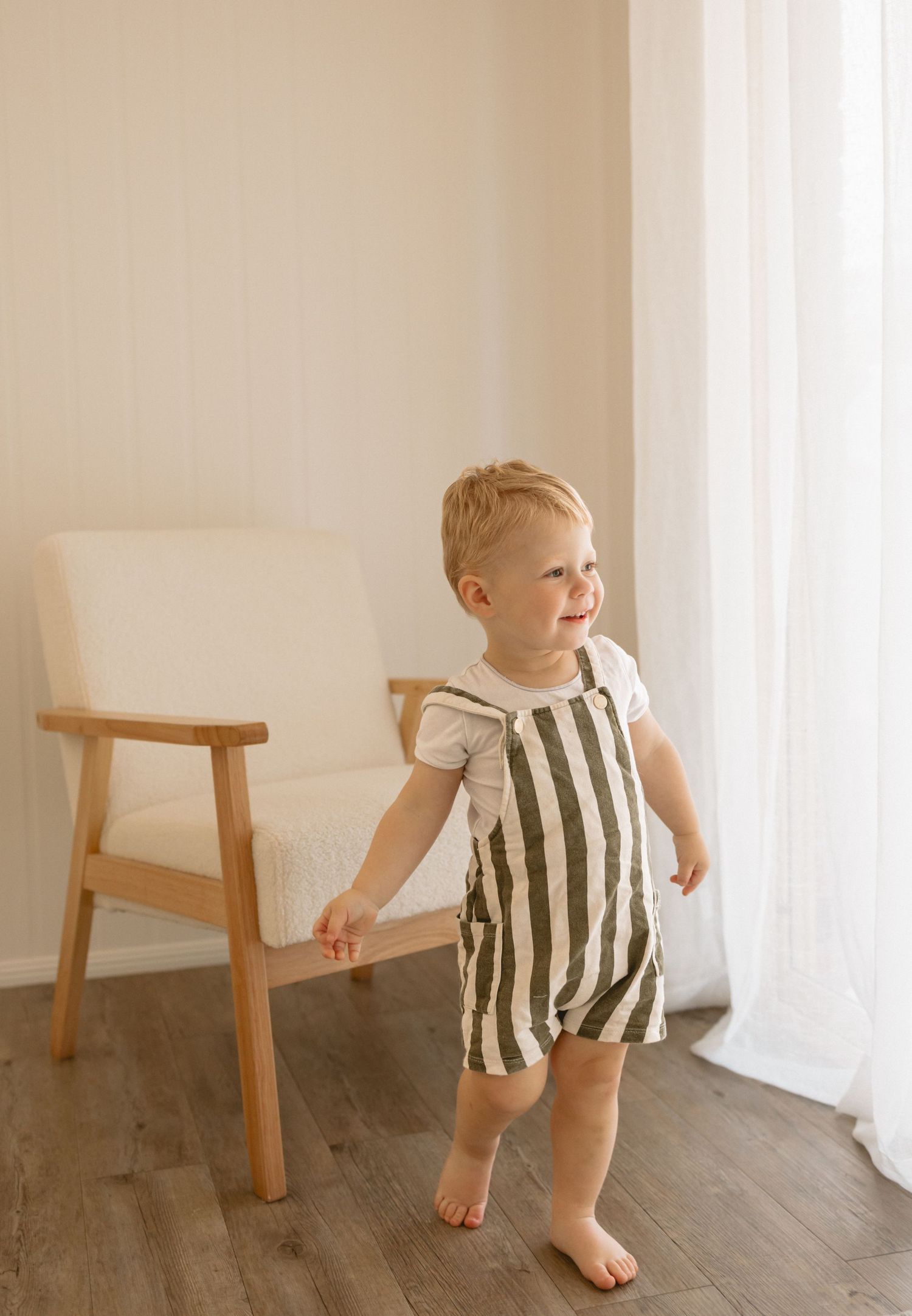 A small child wearing a striped romper stands barefoot next to a wooden stool by a window with sheer curtains.
