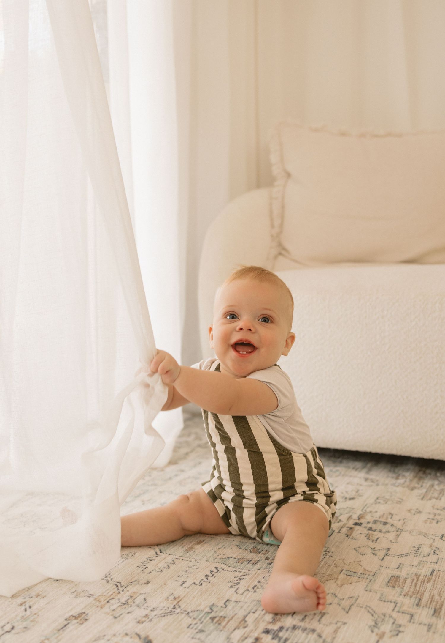 Baby in striped outfit crawls on patterned rug surrounded by white curtains in bright room.