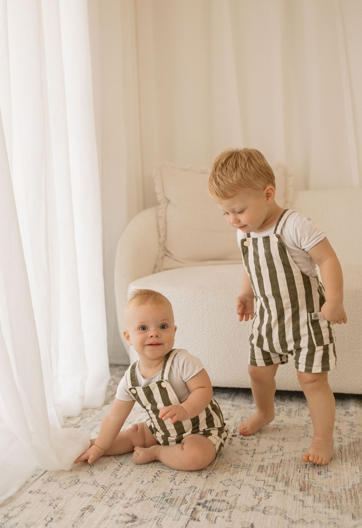 Two young children in matching striped rompers playing together on a light wooden floor by white curtains and sofa.