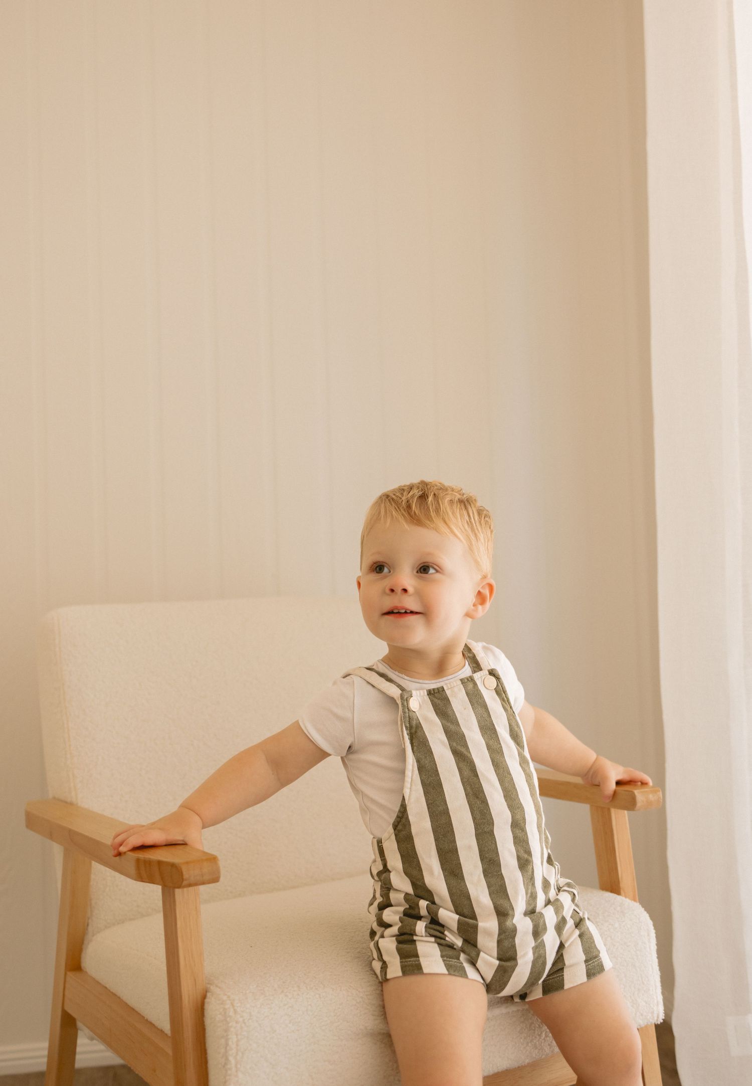 A small child sits on a wooden chair wearing a striped romper against a neutral beige wall.