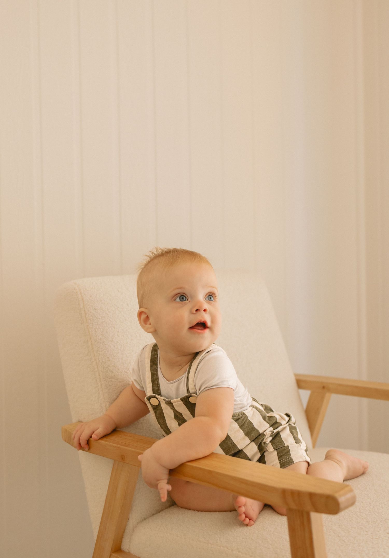 A small child in white t-shirt and striped overalls sits in a wooden armchair against a light-colored wall.