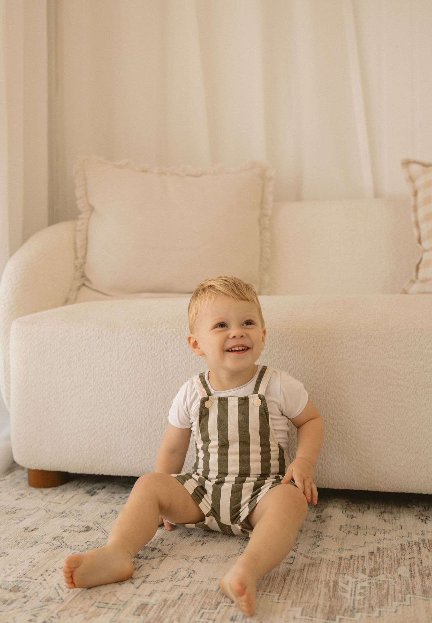 Small child wearing striped overalls and white shirt sits smiling on floor by white couch in minimalist room.