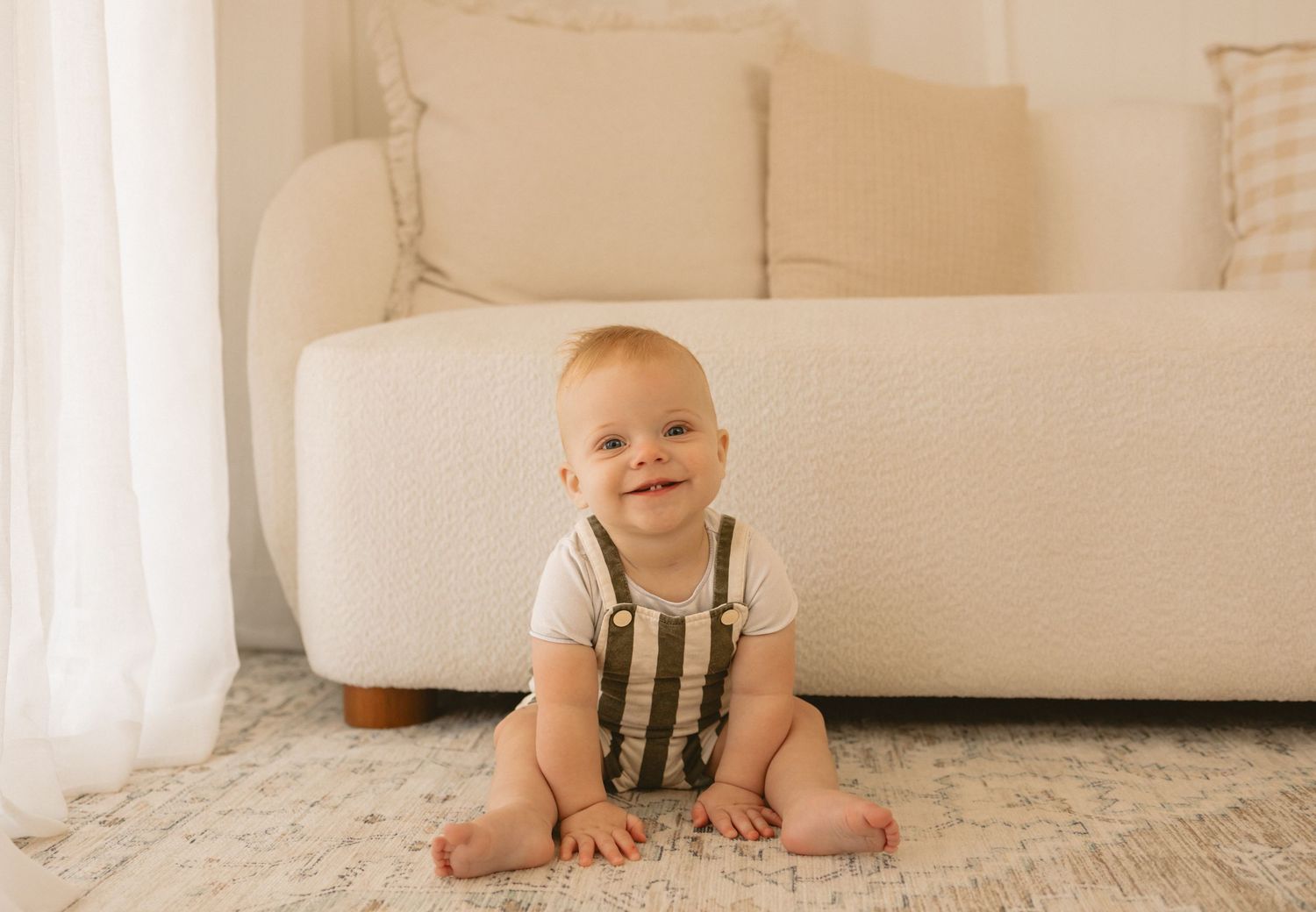 A small baby in a beige onesie crawls on a plush white carpet while playing with toys.