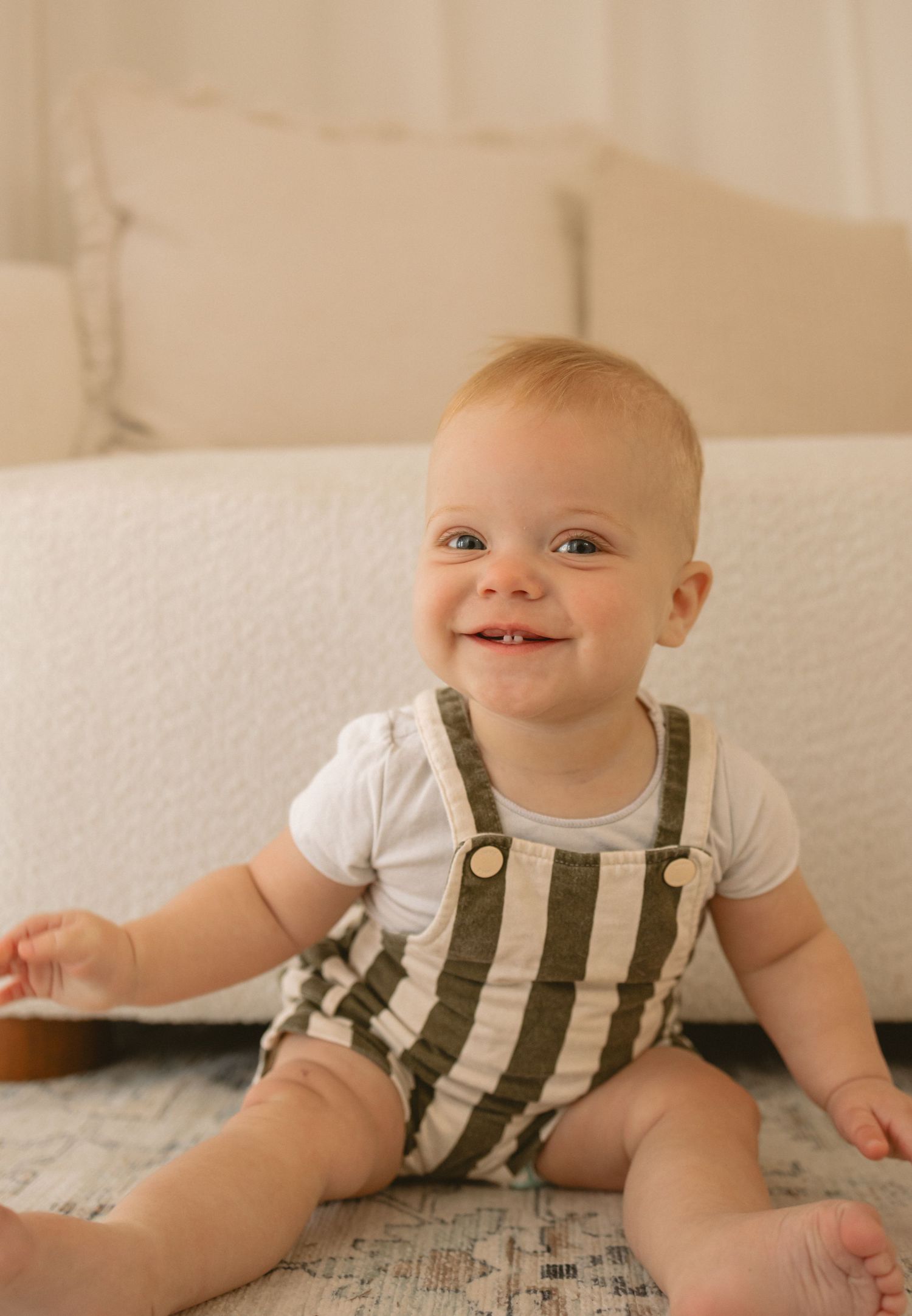 A small child sits on a blanket wearing a white shirt and striped overalls with a joyful expression.