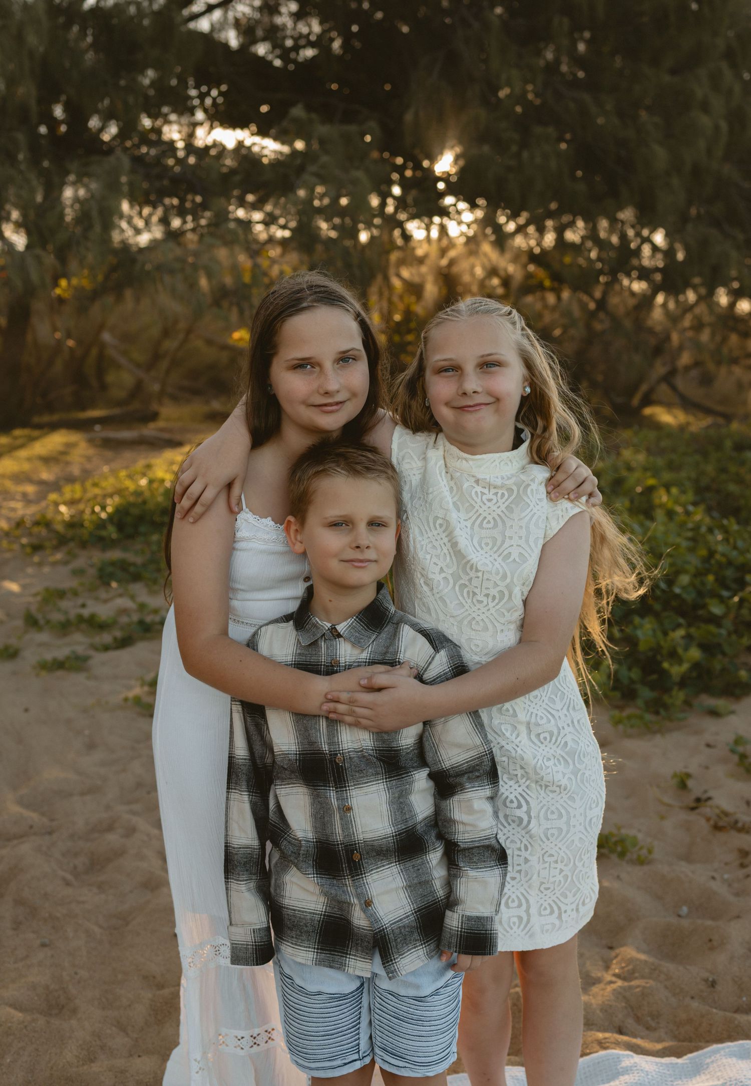 Three siblings embracing outdoors during golden hour on sandy ground with natural backlit lighting.