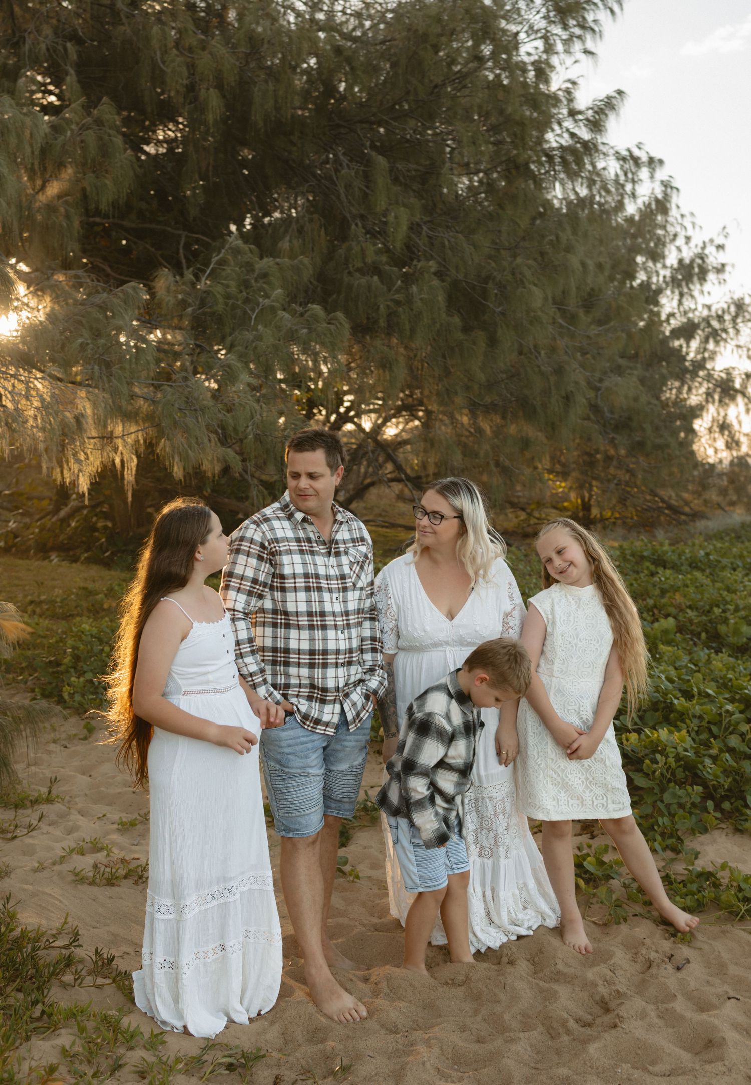 A group wearing white and plaid clothing stands together on a sandy beach at sunset with trees in the background.