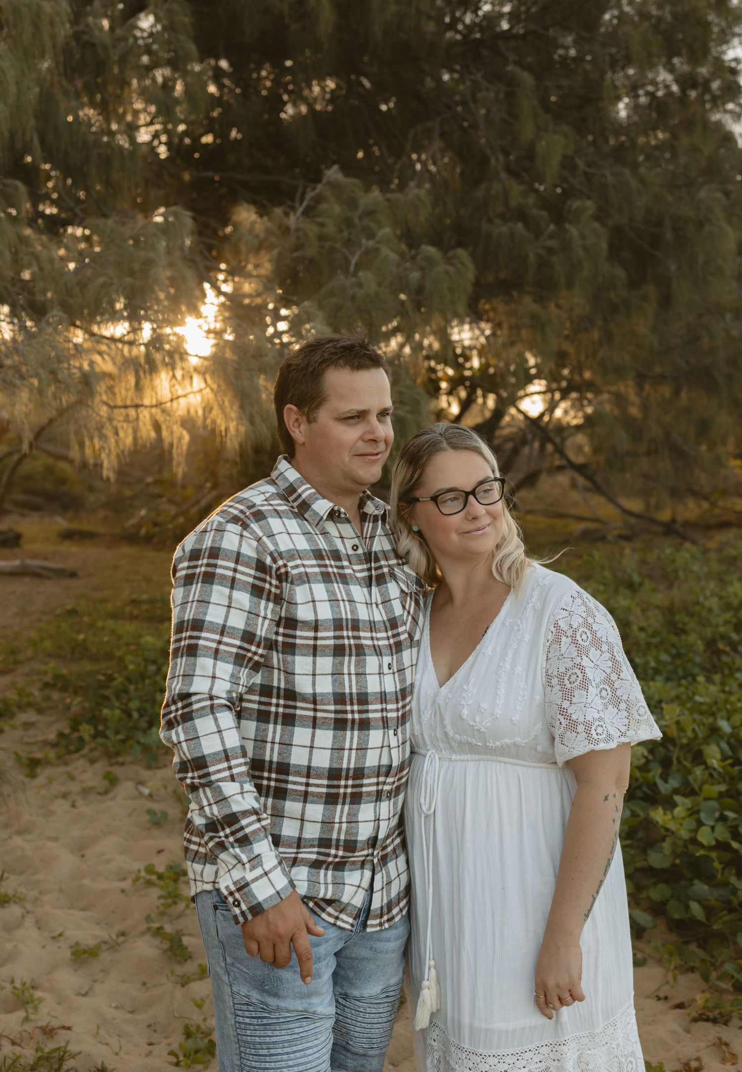 A couple in casual attire shares intimate moments at sunset on a beach surrounded by trees and foliage.
