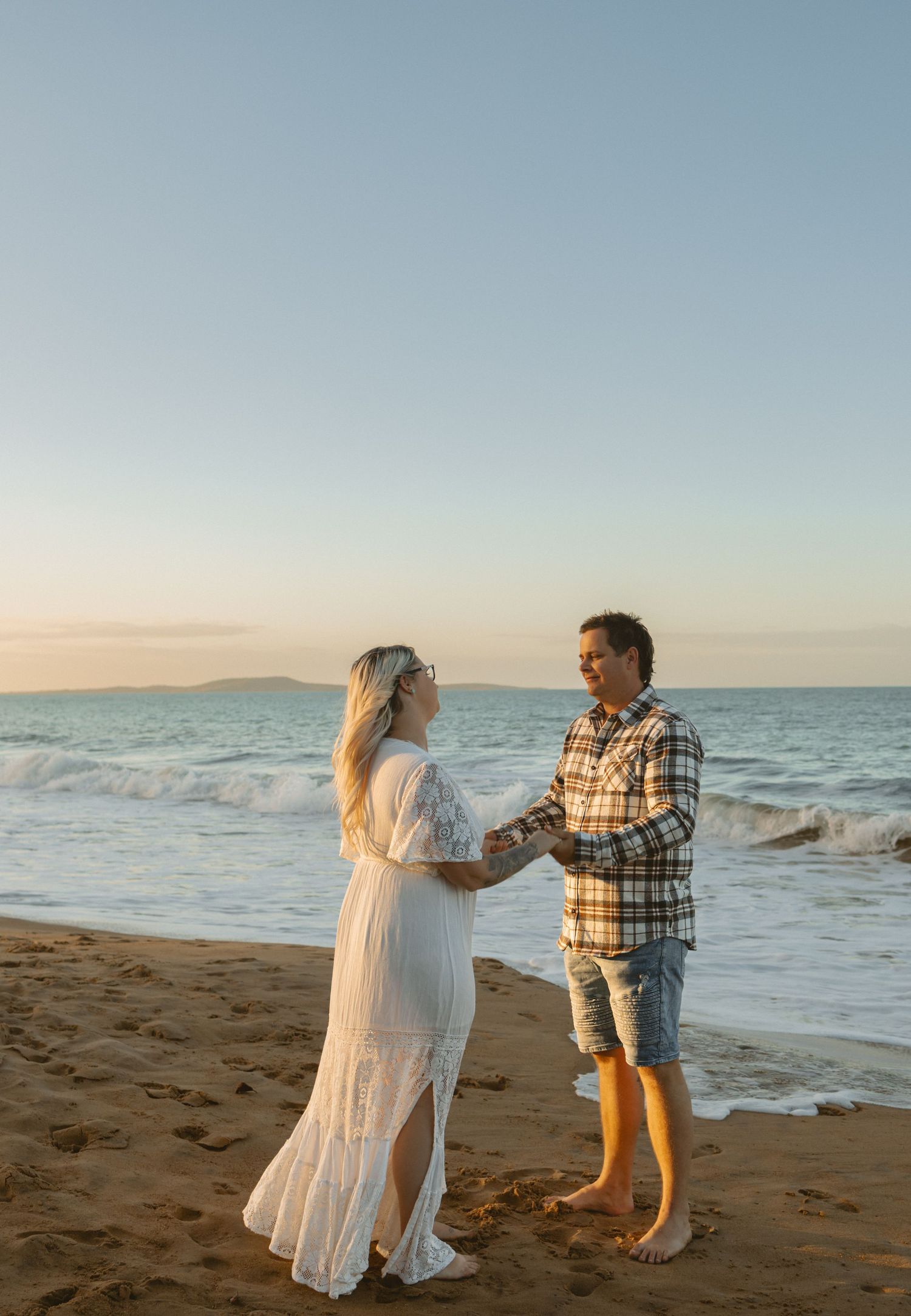 A couple holds hands on a sandy beach at sunset while ocean waves roll in along the shoreline.