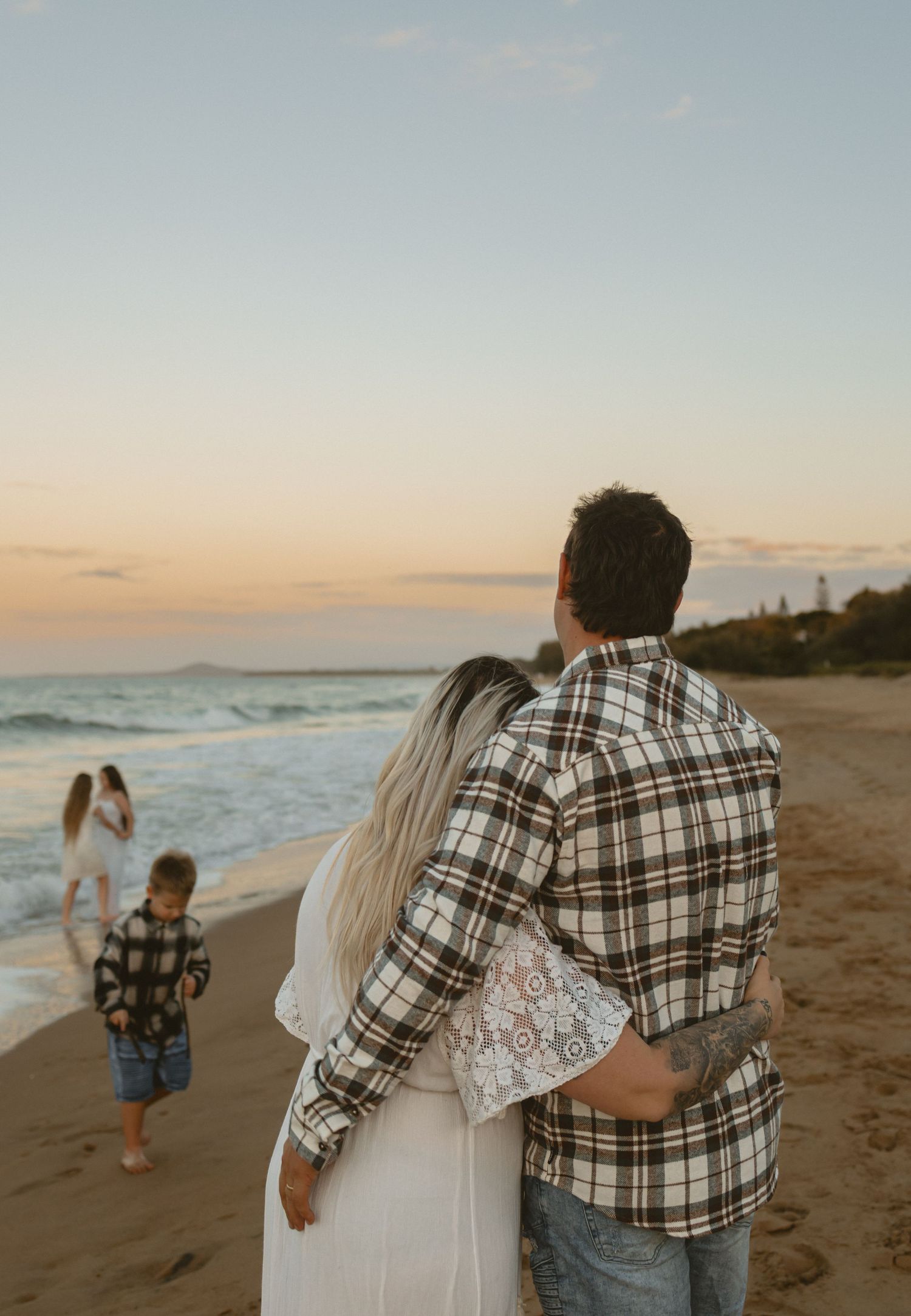 A couple walks along a serene beach at sunset wearing a plaid shirt and white dress, creating romantic silhouettes.