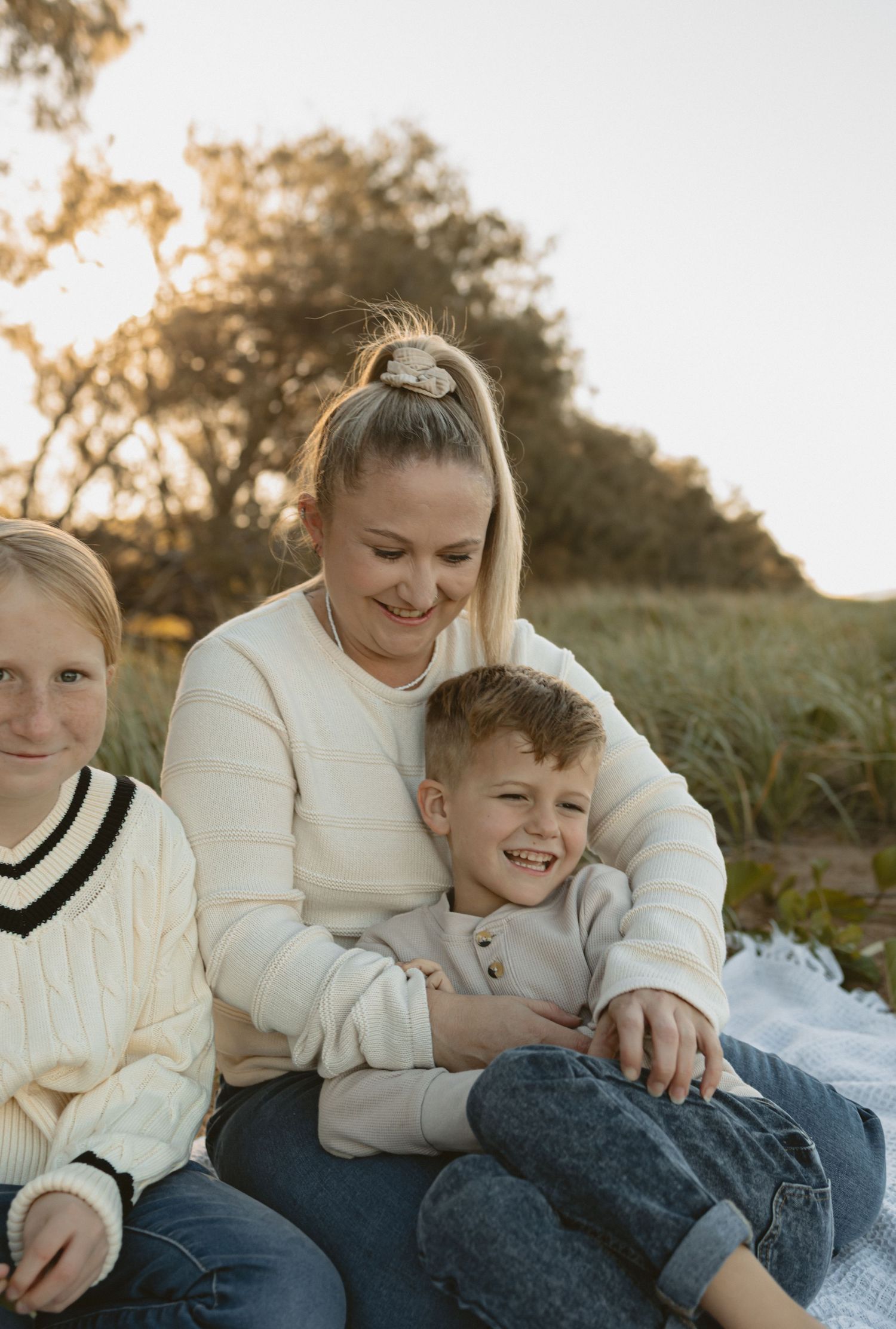 Candid family moment outdoors at sunset, sharing affectionate embraces while wearing cozy white sweaters on a blanket.