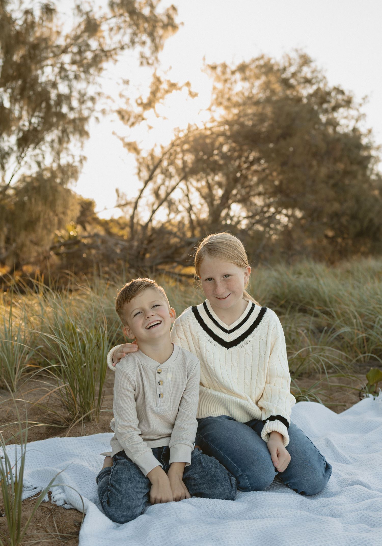 Two children sit together on a white blanket outdoors at sunset, sharing a joyful moment in nature.