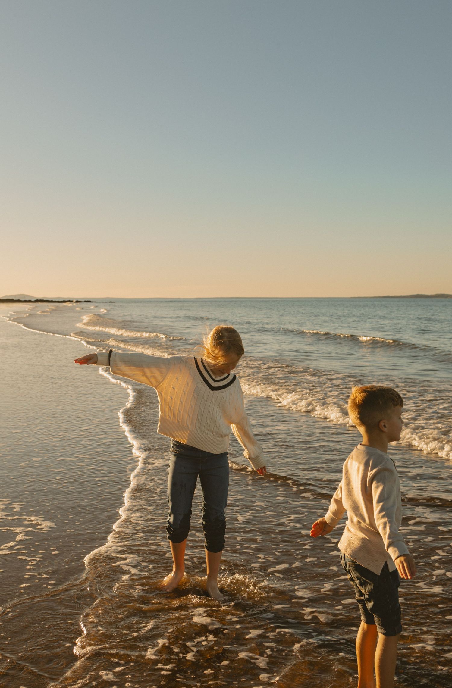 Two young people in sweaters play at the ocean's edge during sunset with gentle waves washing onto the sandy beach.