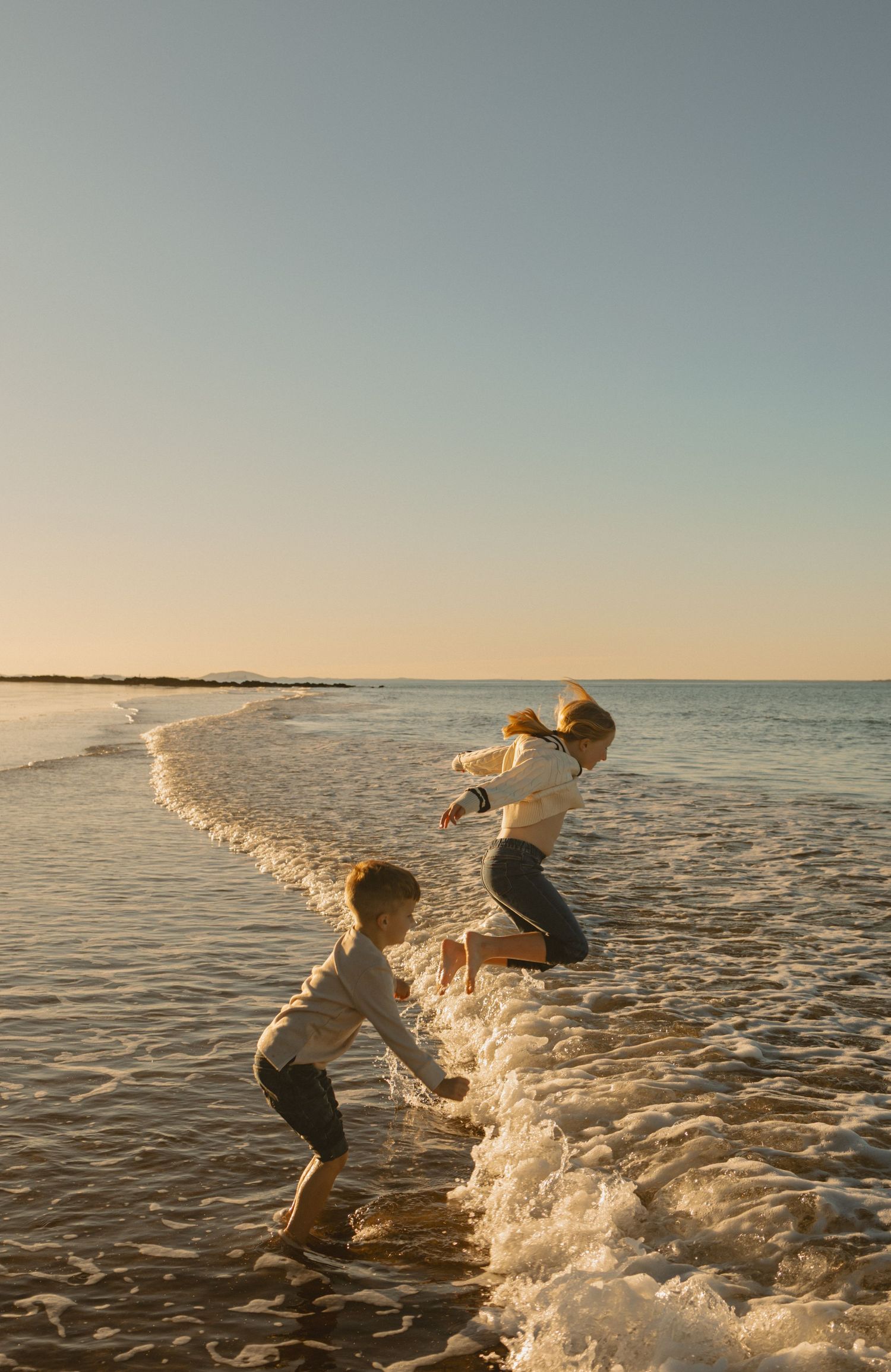 Two people jump playfully through the ocean waves at sunset along a peaceful beach shoreline.