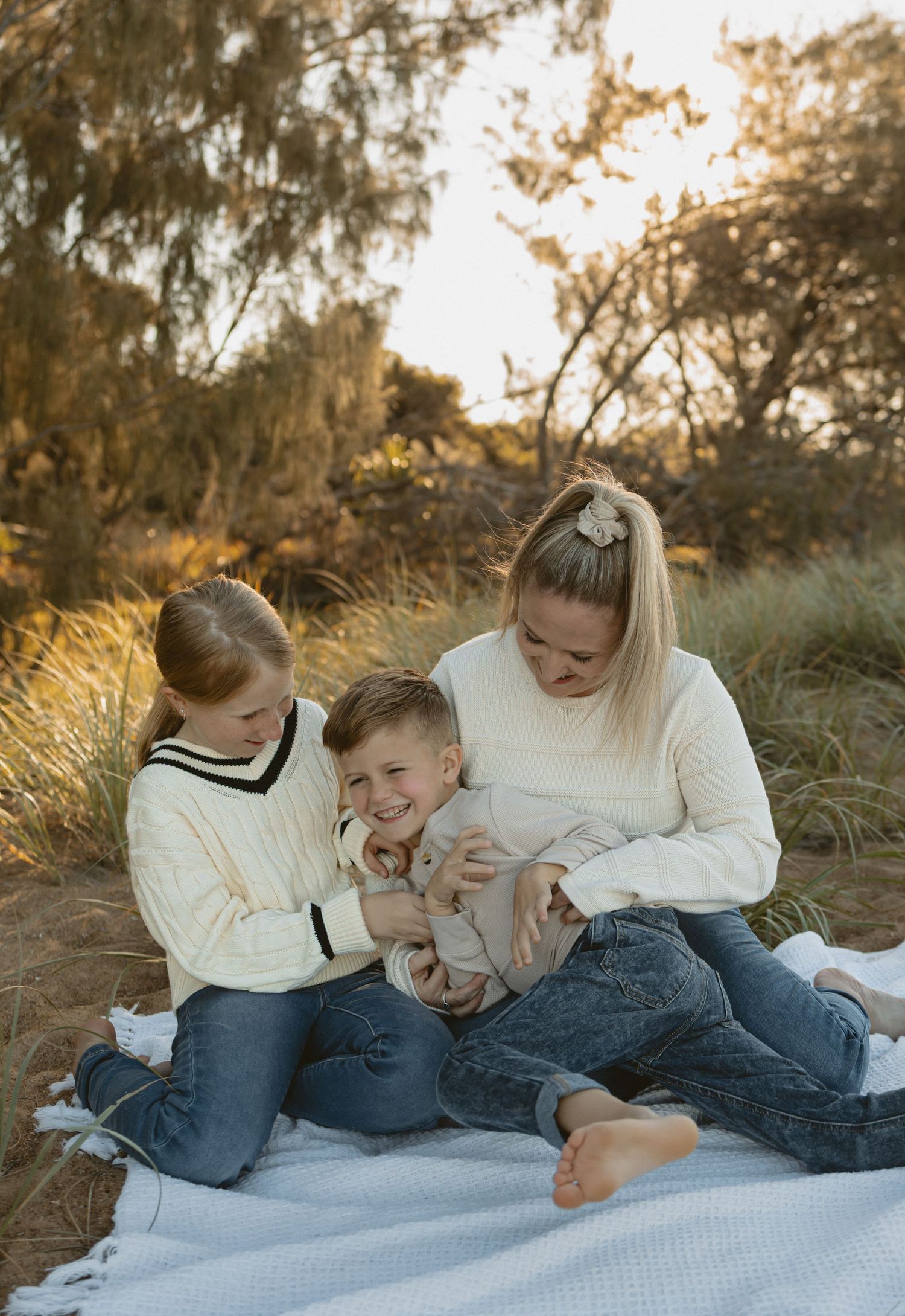 Family members wearing white sweaters cuddle together on a blanket outdoors during golden hour.