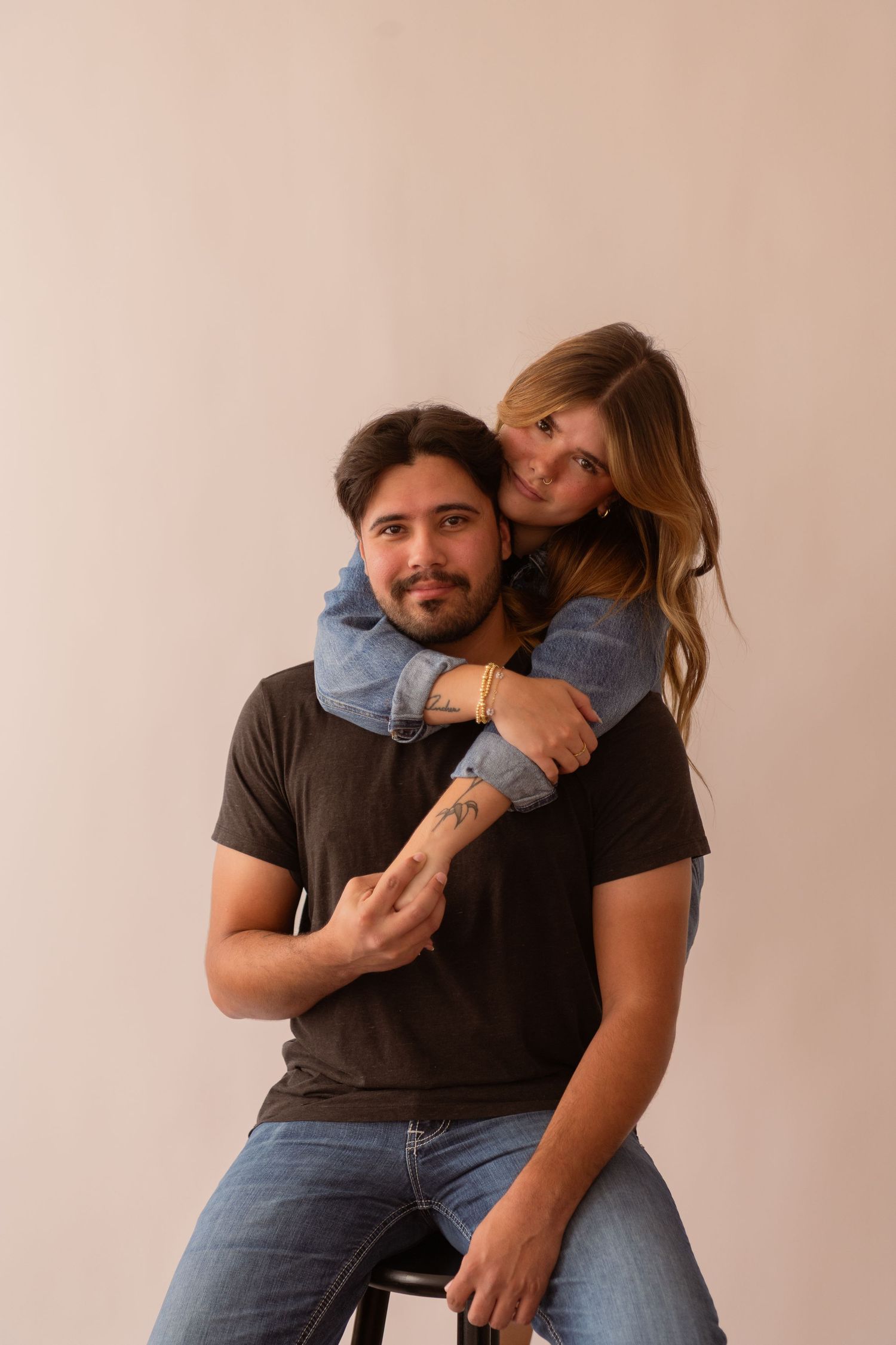 Couple posing in studio, woman in denim jacket hugging man from behind as he sits smiling.