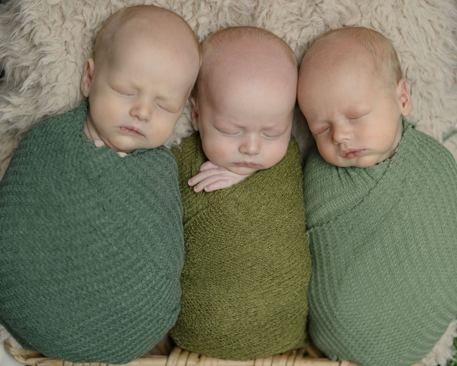 Three sleeping infants snuggled together in green knitted wraps on a textured background.