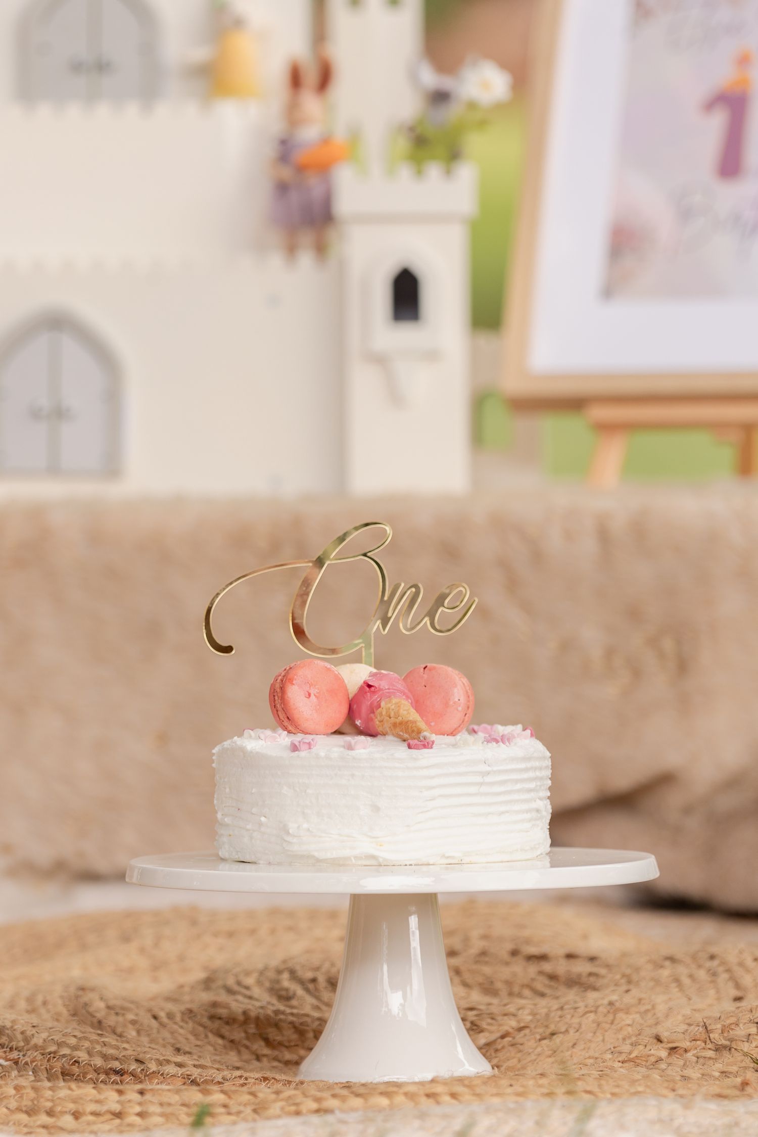 White birthday cake with pink macarons and gold 'one' topper on elegant cake stand.