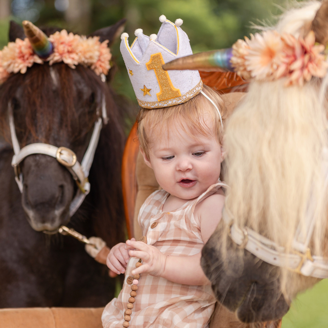A small figure wearing a crown sits between two horses adorned with peach flowers. One Year Old Birthday Party Photoshoot.