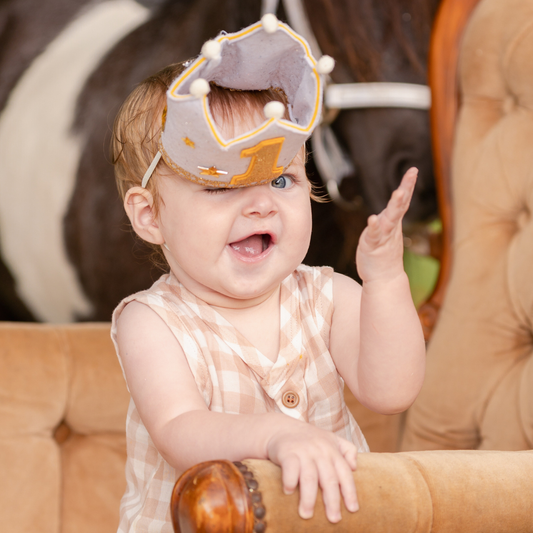 A delightful moment wearing a golden crown while sitting on a tufted chair. One Year Old Birthday Party Photoshoot.