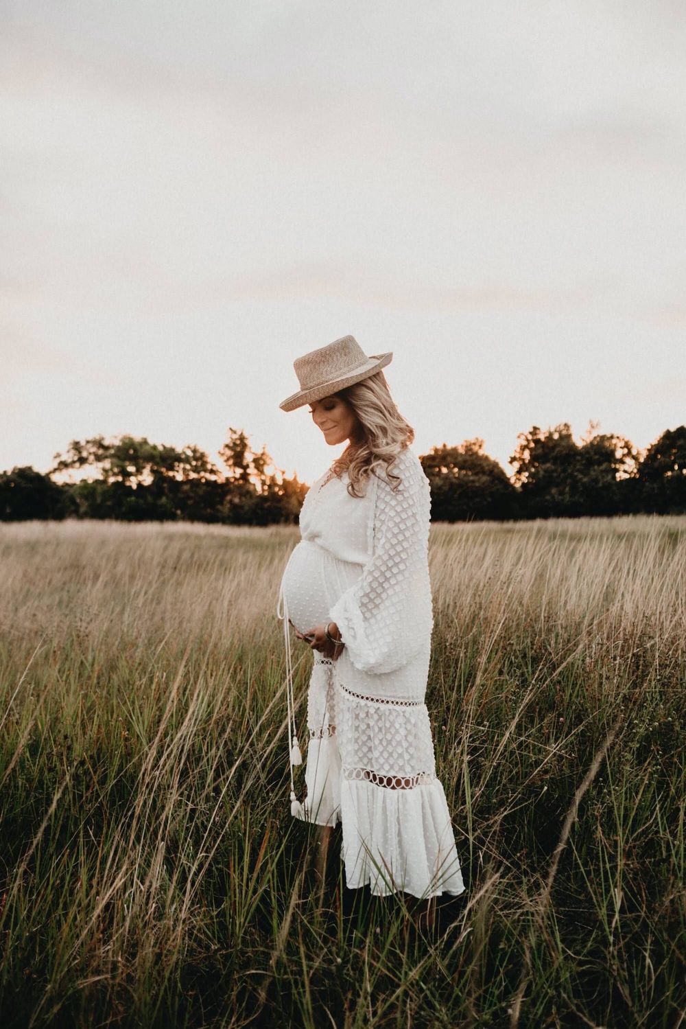 Pregnant woman in white lace dress and hat standing in golden field at sunset.