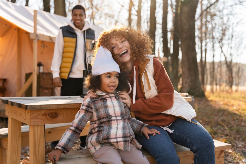 People laughing together and enjoying outdoor camping vibes while sitting at a wooden picnic table near an orange tent.