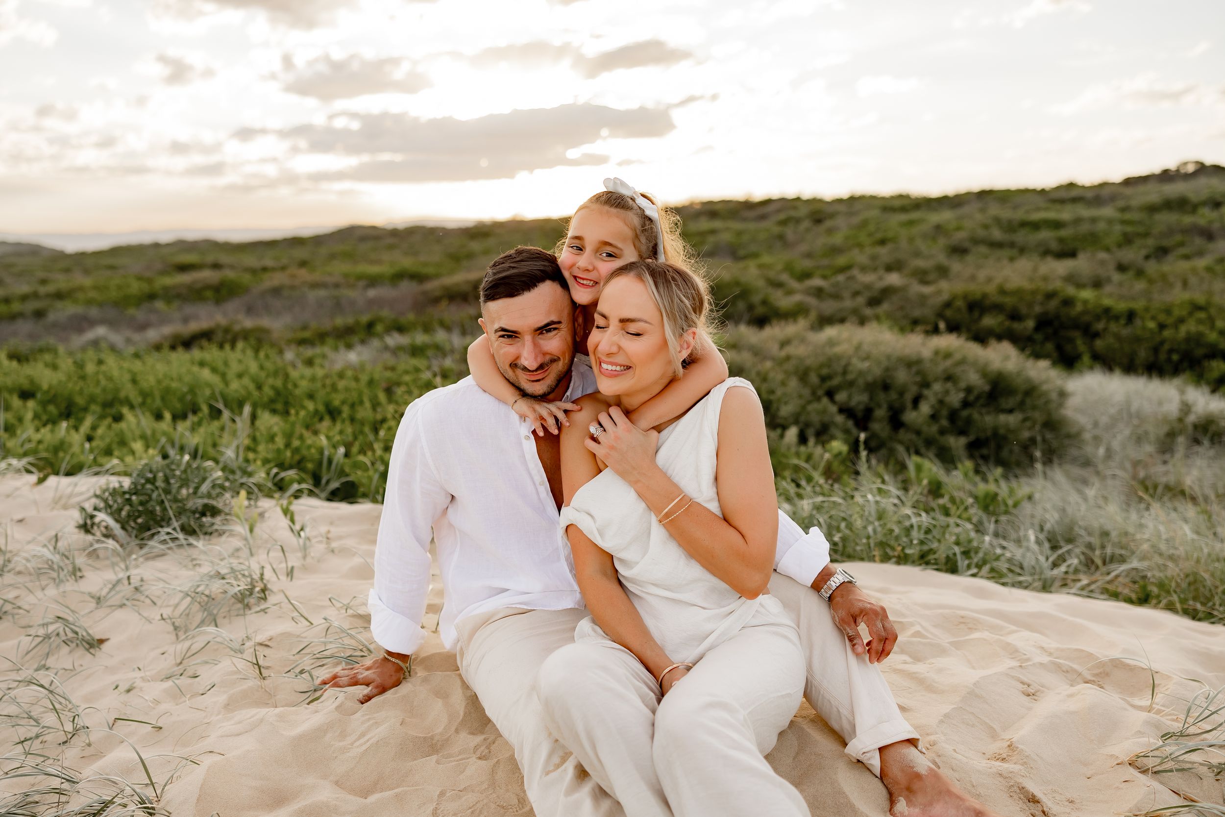 Couple in white clothing embraces while sitting on sandy dunes at sunset.