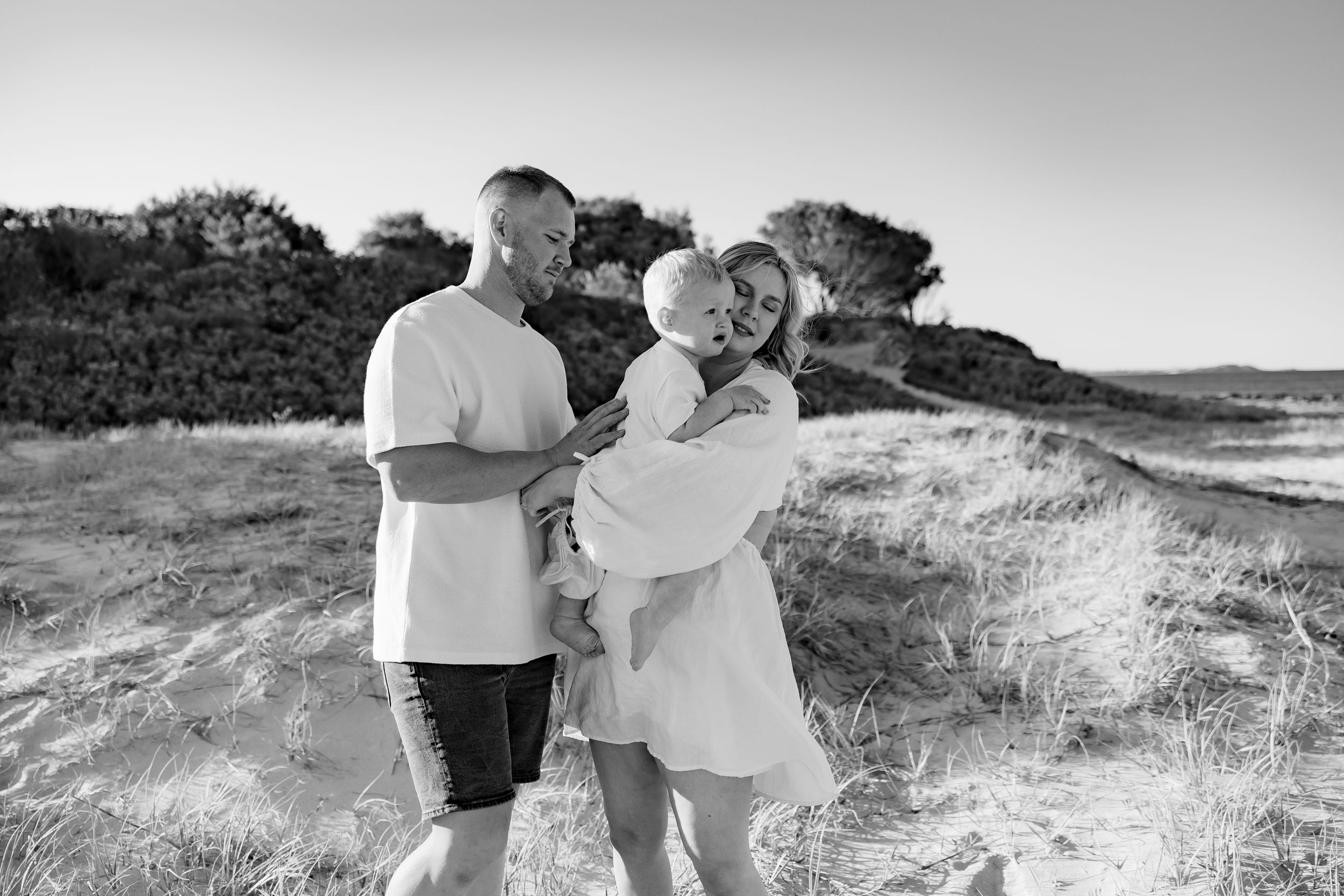 Black and white portrait of a family sharing a tender moment on coastal dunes.