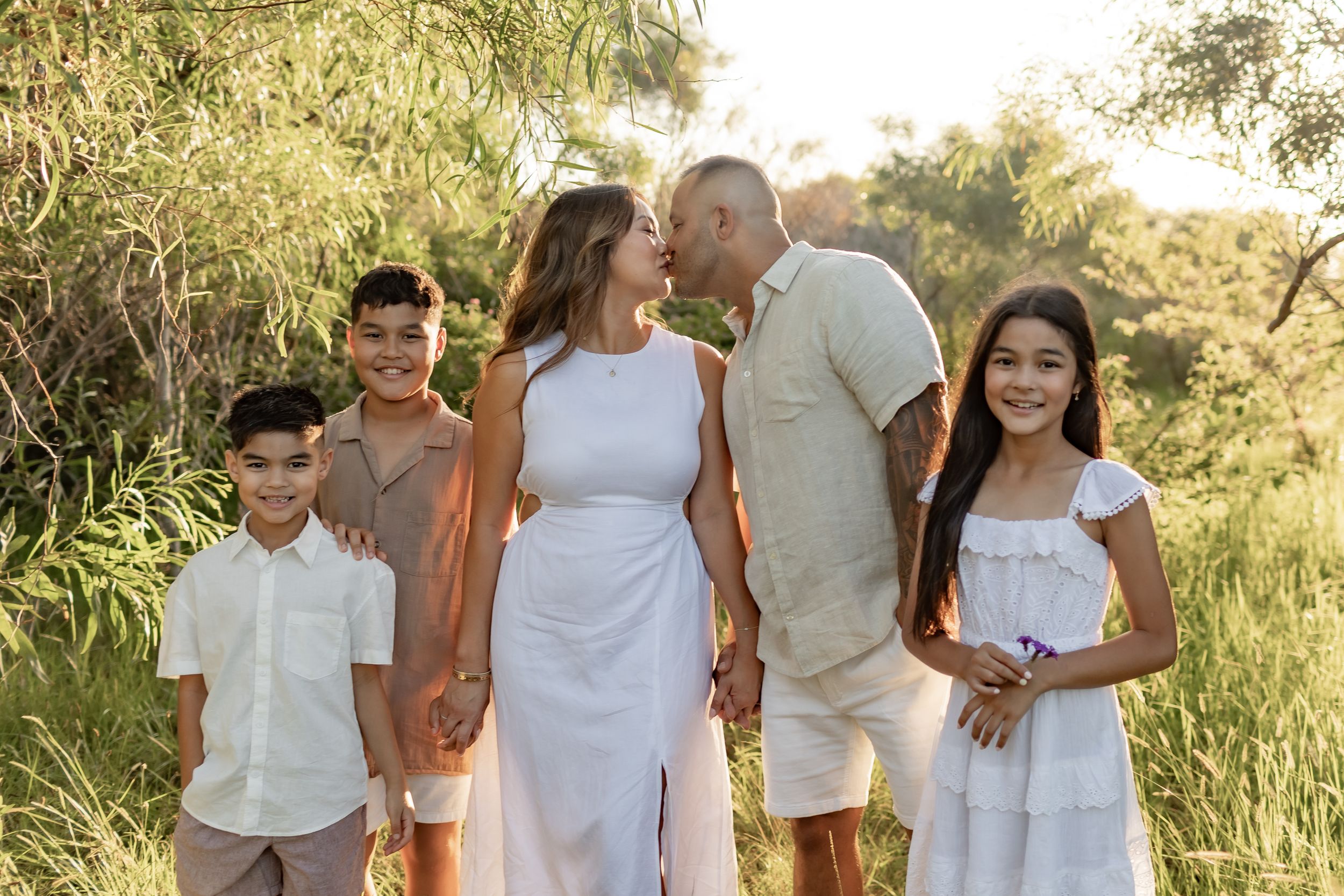 A family wearing coordinated white outfits poses together in a natural outdoor setting at sunset.