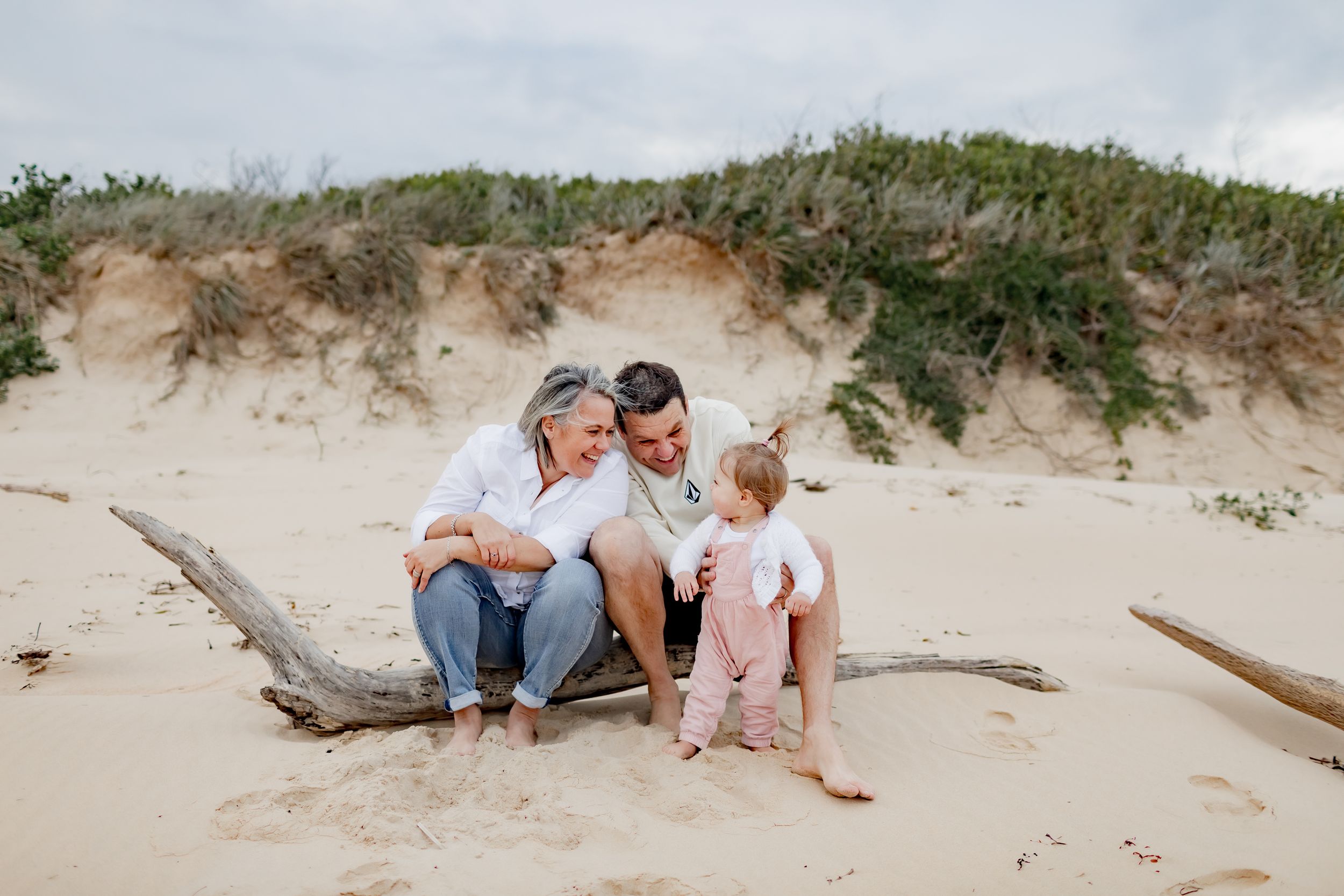 Family sitting together on driftwood at a scenic beach with sand dunes.
