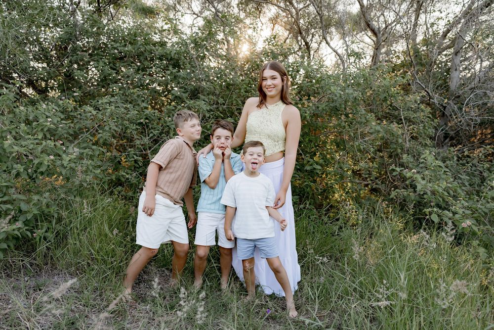 A family poses together outdoors in casual white outfits against a natural green foliage background at sunset.