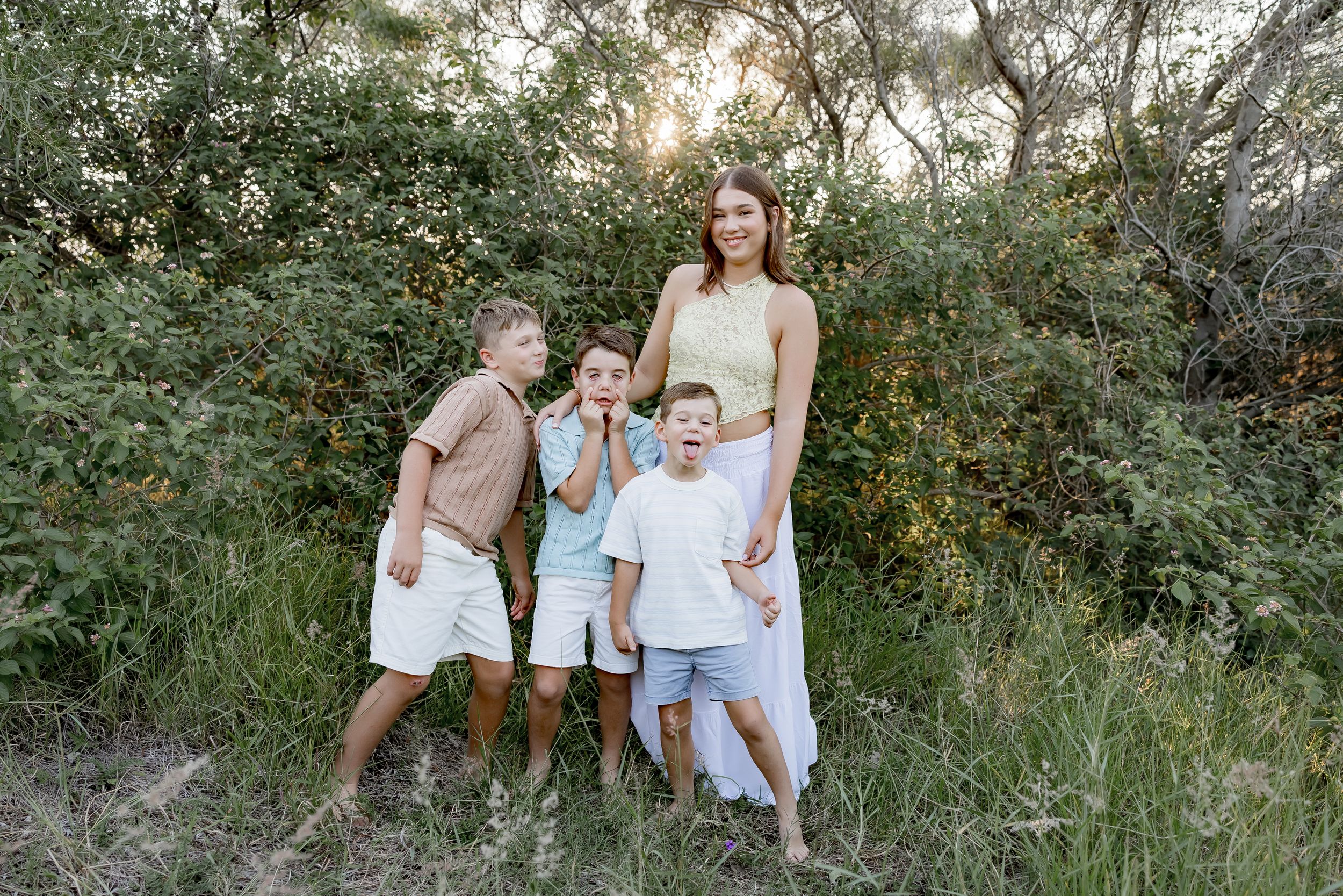 A family poses together outdoors in casual white outfits against a natural green foliage background at sunset.