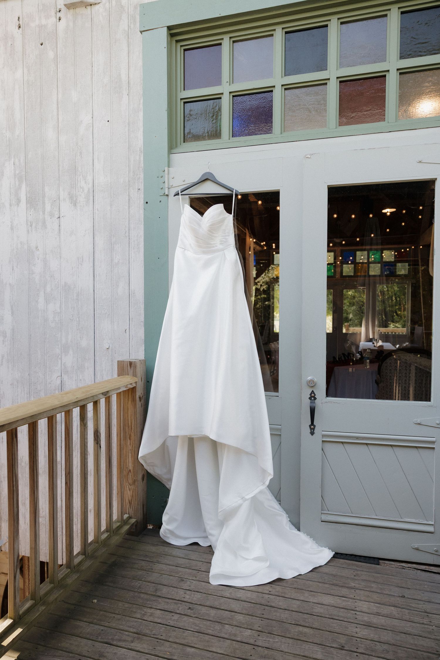 A white wedding dress hangs on display outside a green-framed door on a wooden deck.