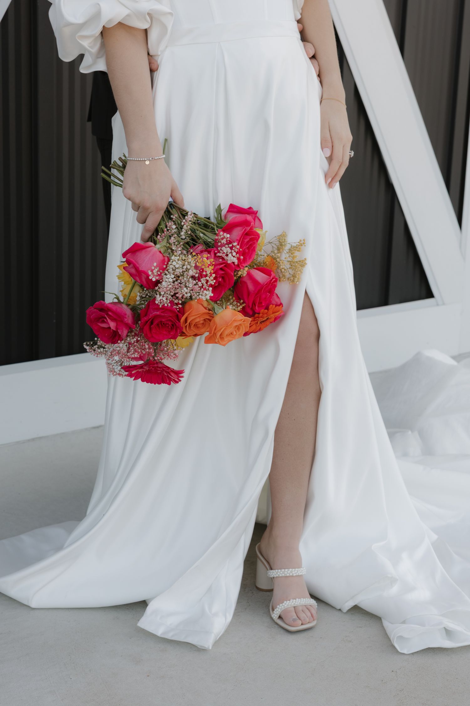 Close-up of bride in flowing white dress with slit holding vibrant pink and orange bouquet with baby's breath details.
