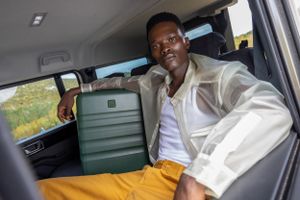 Person sitting in car with green Antler luggage wearing white shirt and yellow pants.