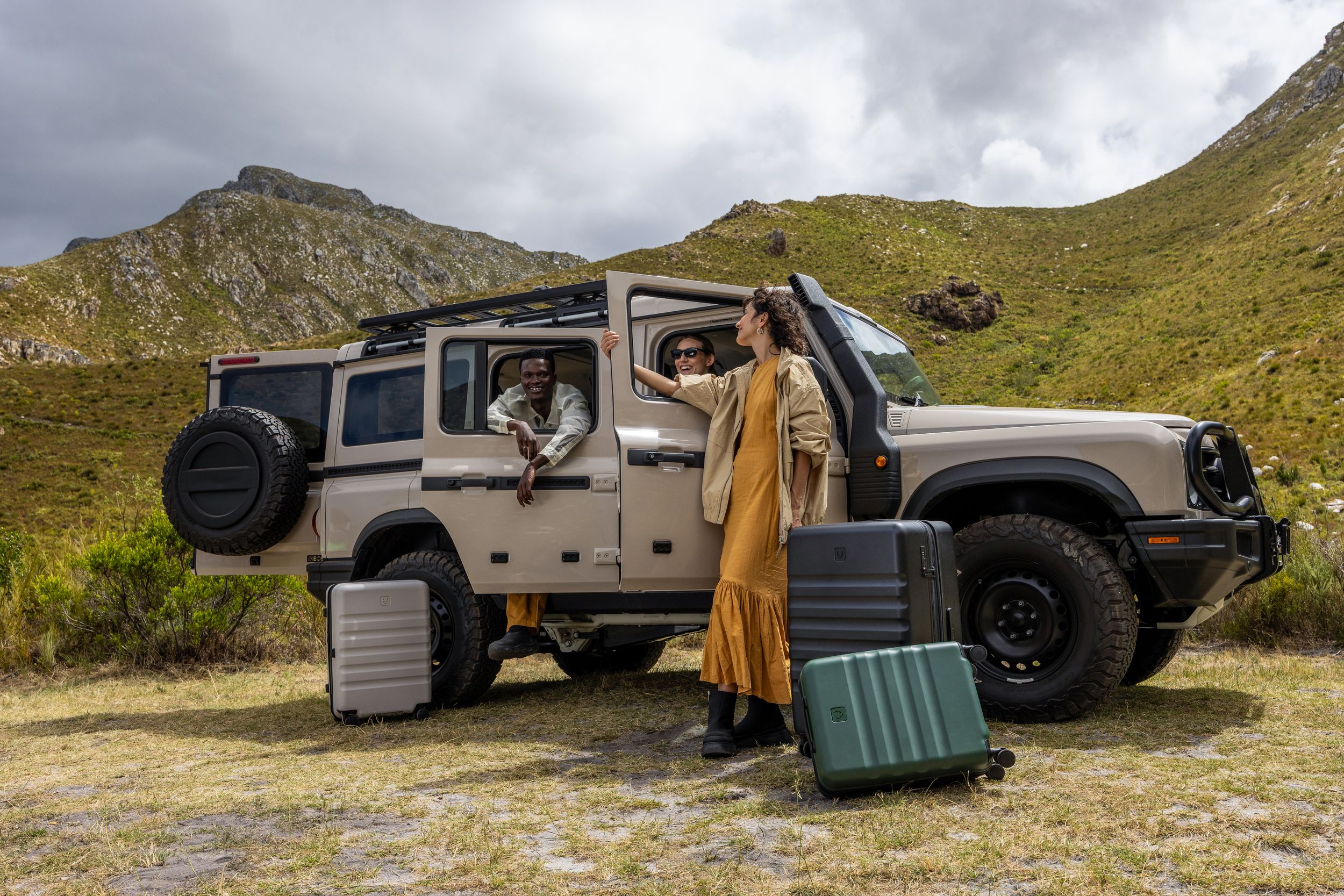 Adventure travelers with suitcases pose next to a rugged off-road vehicle in mountain terrain.