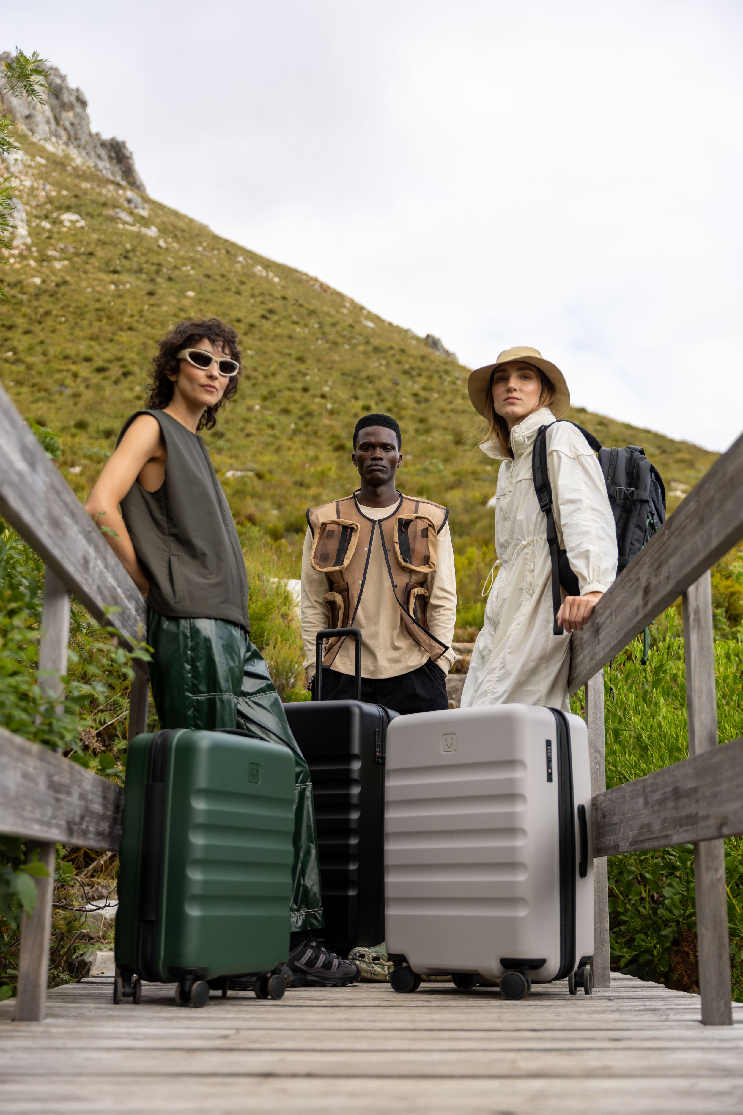 Three travelers with luggage standing on wooden platform against mountain backdrop.