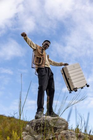Person standing triumphantly on rock holding silver suitcase against blue sky.