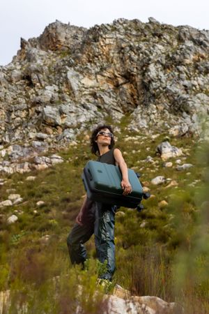 Person holding green suitcase while standing on rocky outcrop in mountainous terrain.