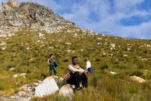 Travelers with luggage hiking on a rocky mountain trail against a blue sky background.