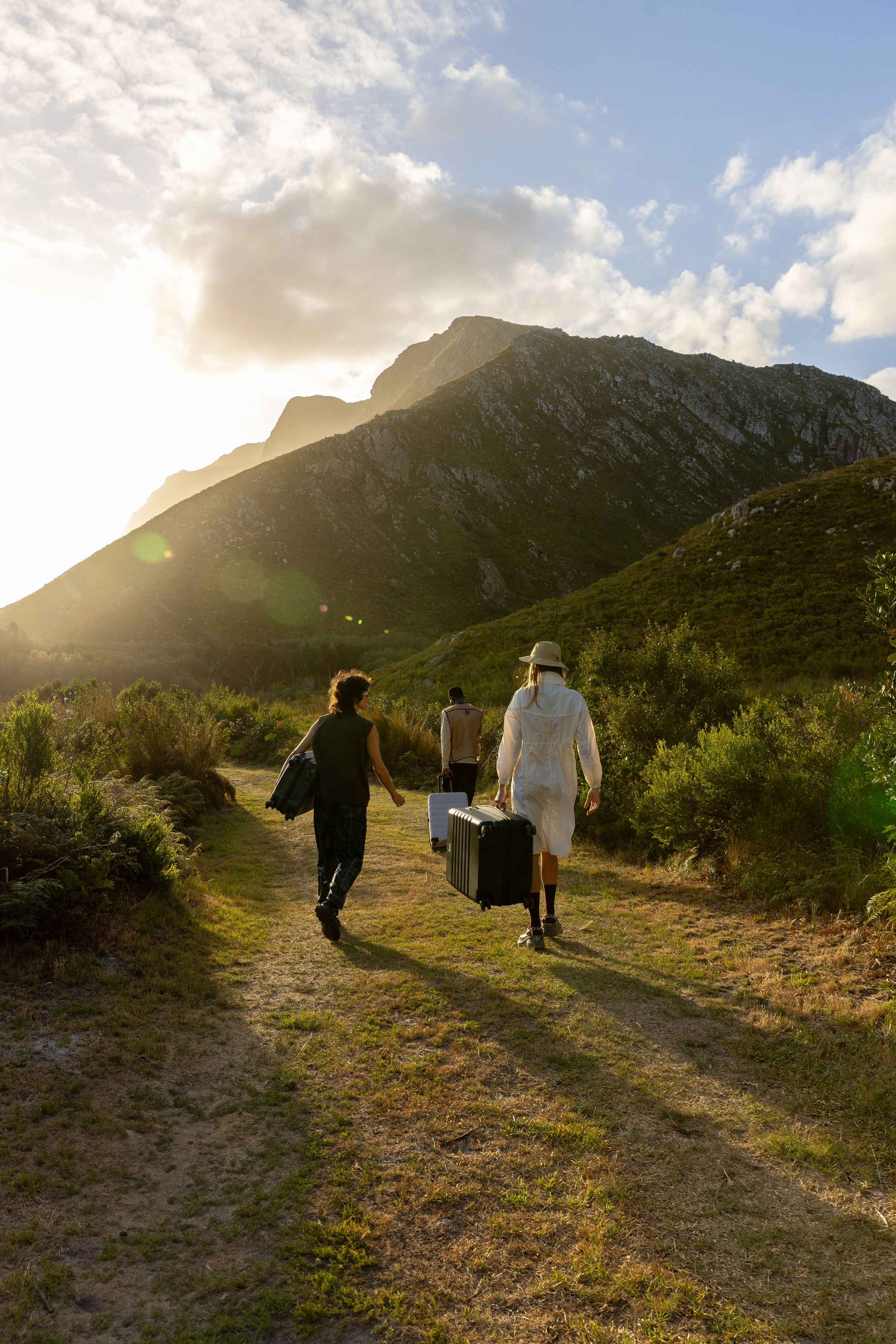 Travelers walking along mountain path with luggage during golden sunset.