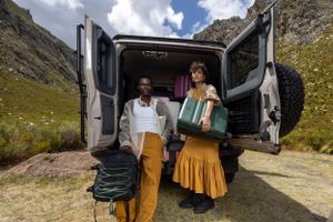 Two travelers stand at the back of a van with luggage against a mountainous backdrop.