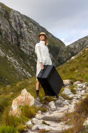 Traveler with suitcase poses on rocky mountain path in scenic landscape.