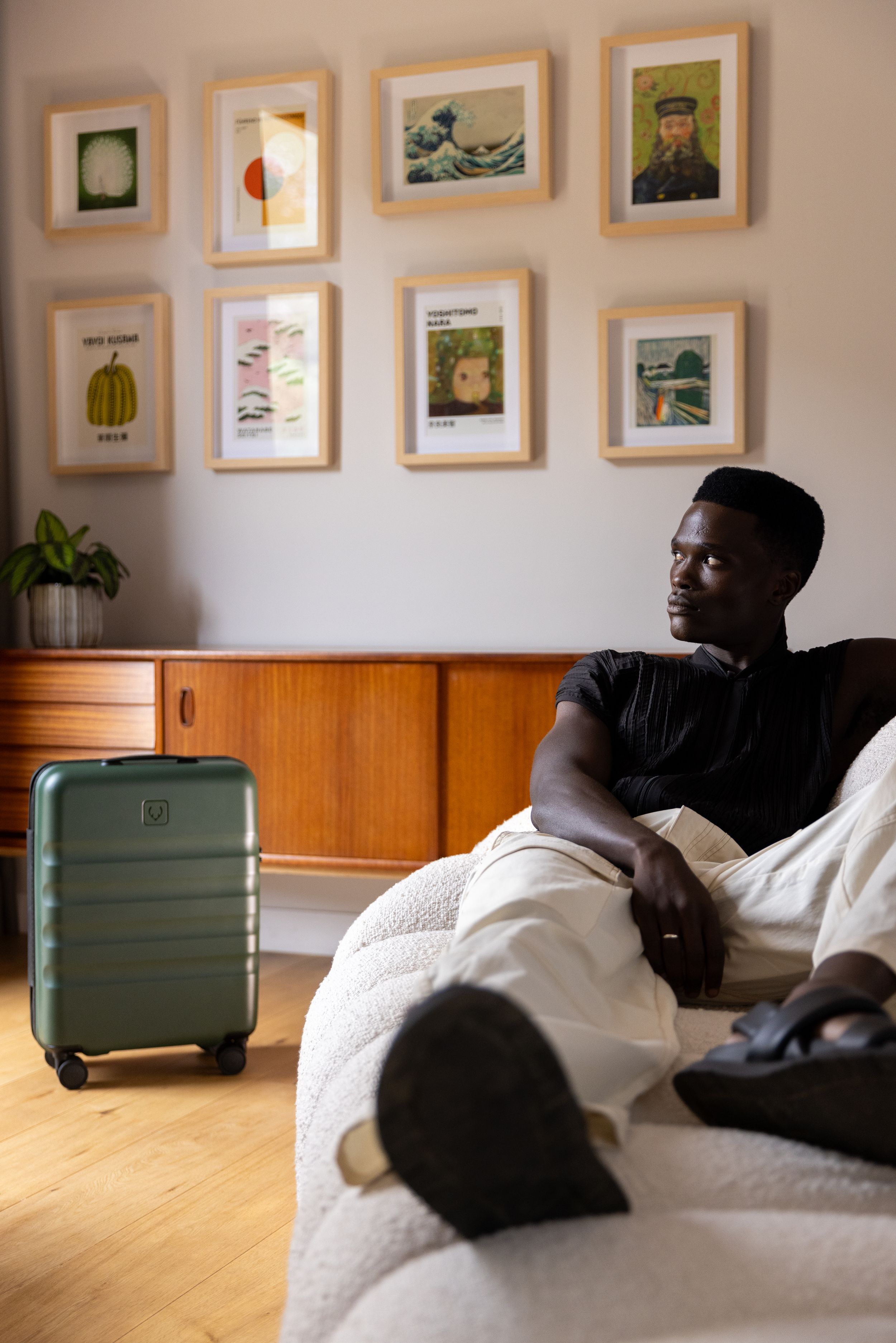 Person relaxing on white couch with green suitcase in room decorated with framed artwork.