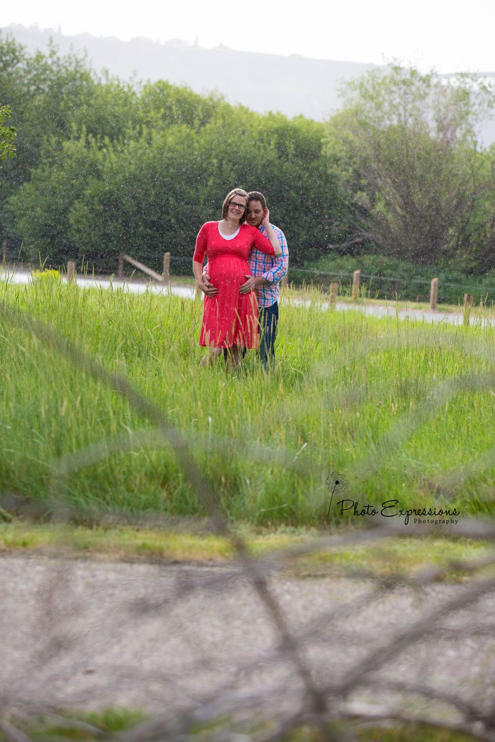 Couple posing together in a green meadow at sunset with trees in the background.