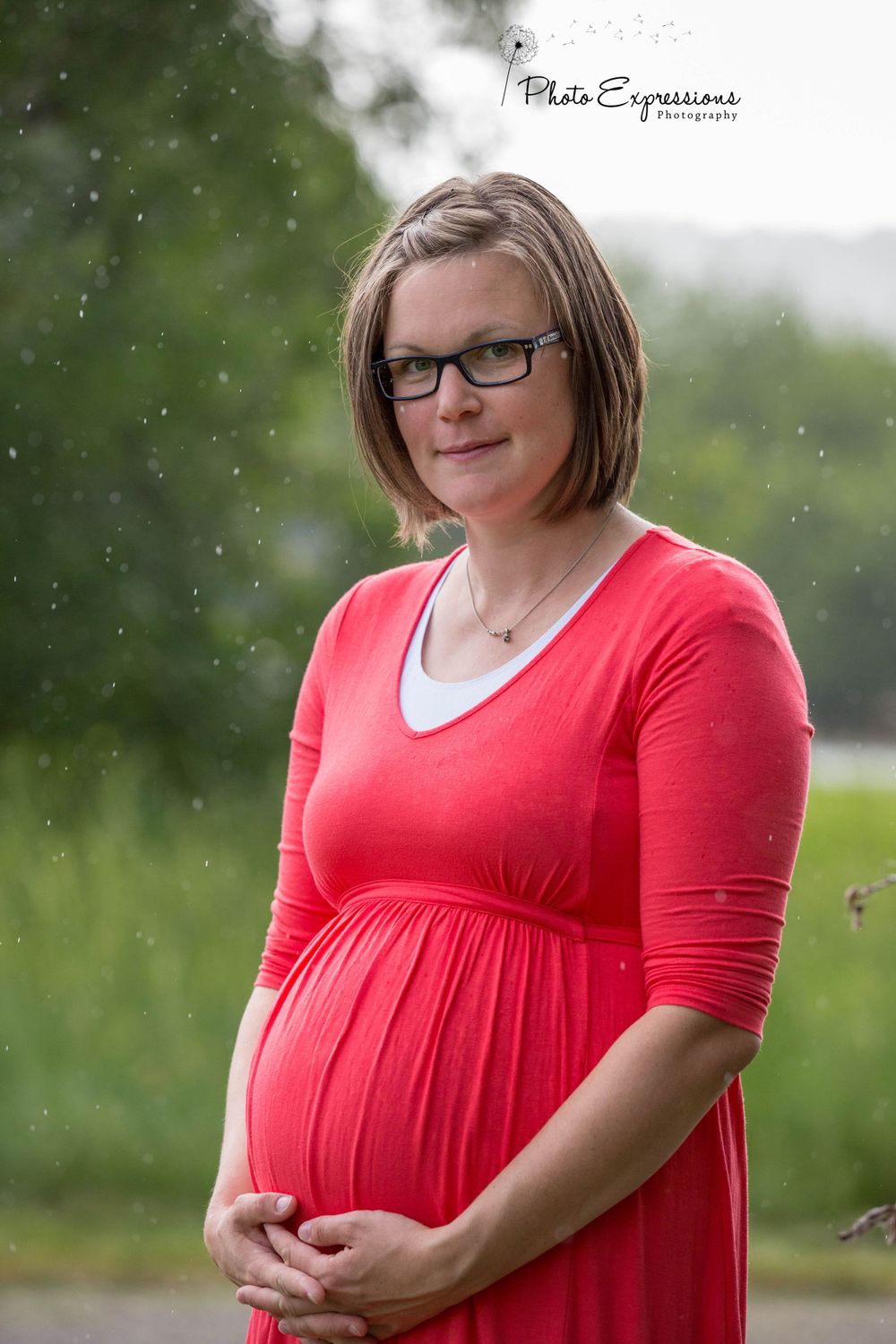 Woman in red dress poses for maternity photos with natural light and rain effects.