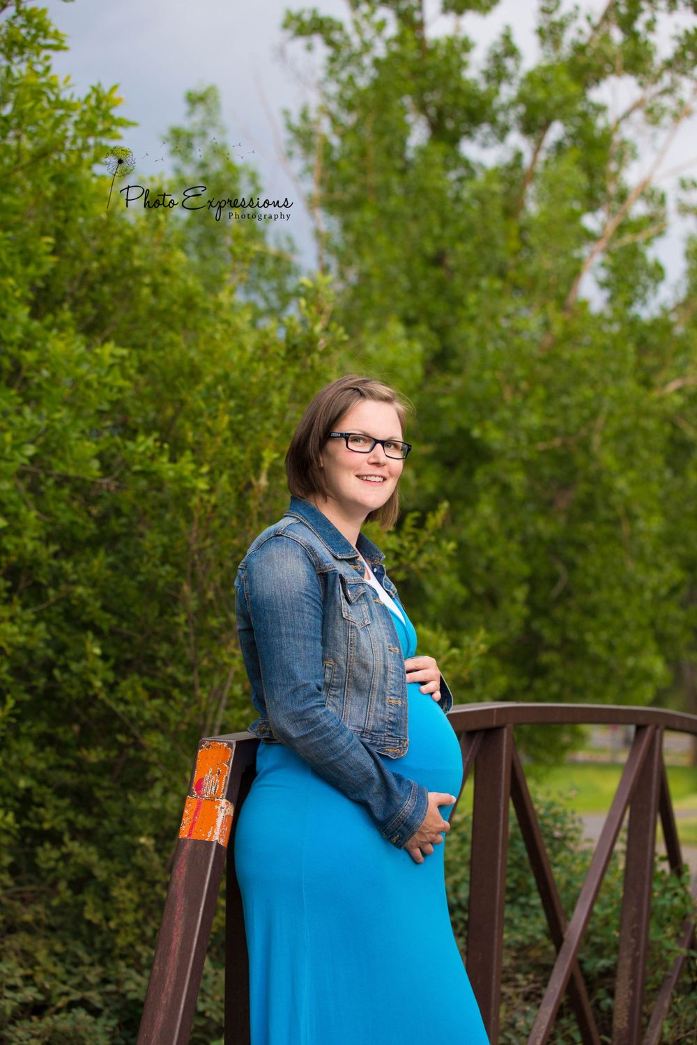 Person in blue dress and denim jacket posing on wooden bridge surrounded by greenery.