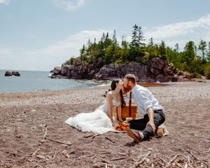 A romantic picnic scene on a rocky beach with dramatic cliffs and evergreen trees in the background.